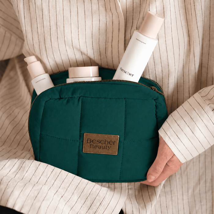 A dark teal quilted cosmetic pouch with a gold-tone zipper and a brown leather patch reading 'Bescher Beauty' is held in a person's hand. The pouch is filled with white skincare bottles, and the person is wearing a cream-colored pinstriped shirt.