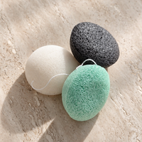 Three natural Konjac Sponges in white, dark gray, and mint green, arranged on a textured beige stone surface with soft leaf shadows.